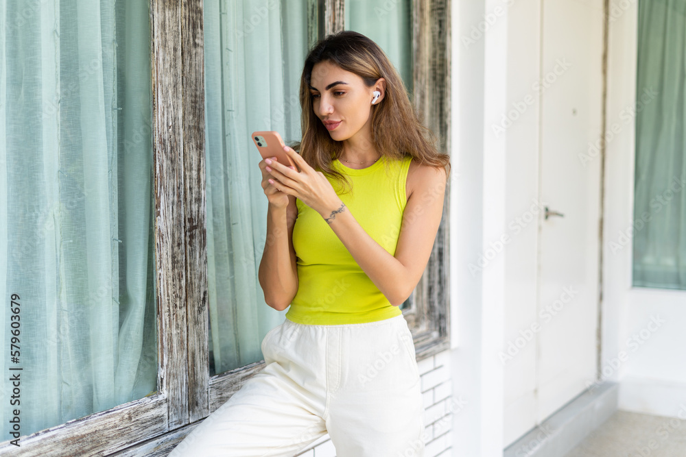Fototapeta premium Welcome. Portrait of cheerful woman standing at modern villa with smartphone and headphones holding phone with happy thoughtful smile