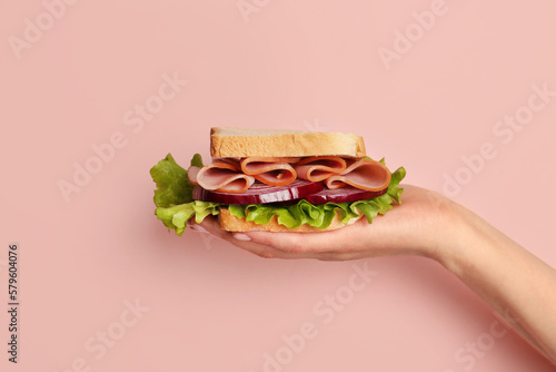 Female hand holding delicious ham sandwich on pink background