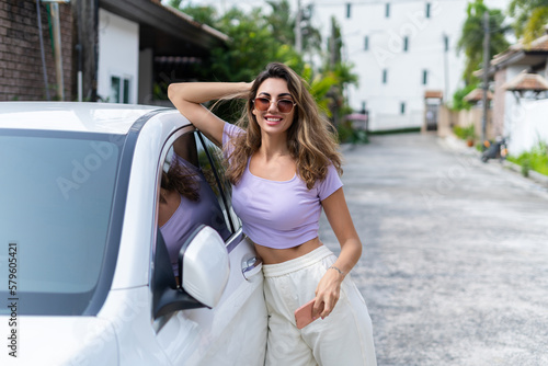 Successful smiling fit tanned attractive woman in casual wear is using her smart phone while standing near luxury modern car outdoors.