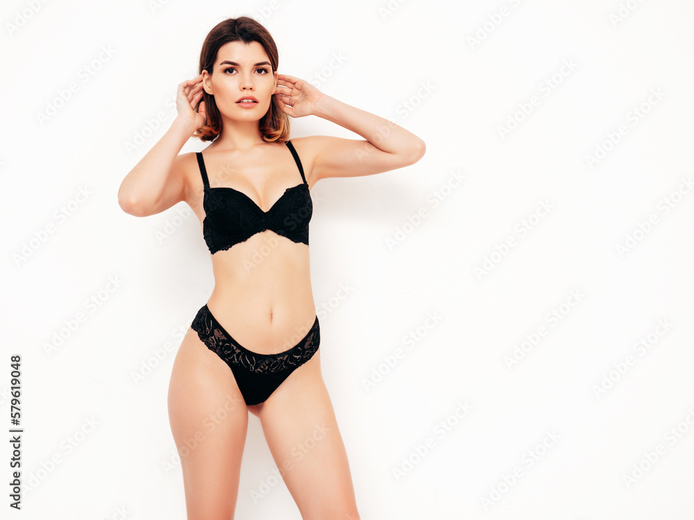 Fototapeta premium Young beautiful sexy woman in black lace lingerie. Carefree smiling model wearing pure underwear. Hot brunette posing near white wall in studio. Perfect body. Slim and fit. Isolated
