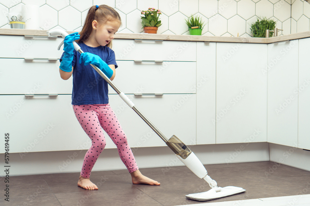 Pretty little girl in household gloves wash floor with modern mop with removable brush near kitchen set. Full length photo of child tidying up kitchen side view. Home wet cleaning concept. Pretty little girl in household gloves wash floor with modern mop with removable brush near kitchen set. Full length photo of child tidying up kitchen side view. Home wet cleaning concept.