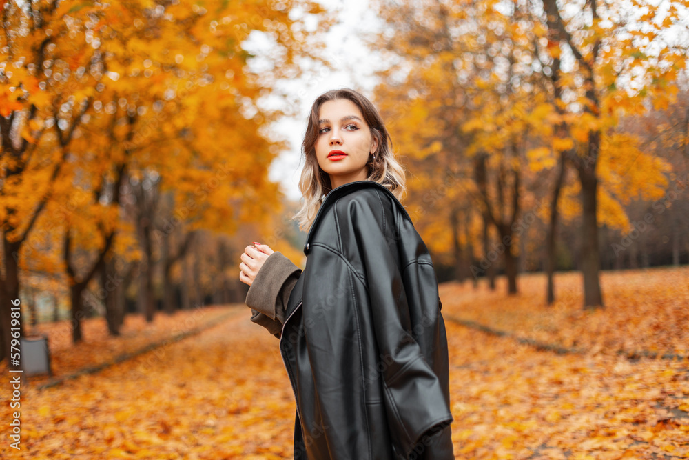 Fashion beautiful trendy girl model with red lips in stylish leather black jacket and pullover walks in amazing golden park with yellow leaves