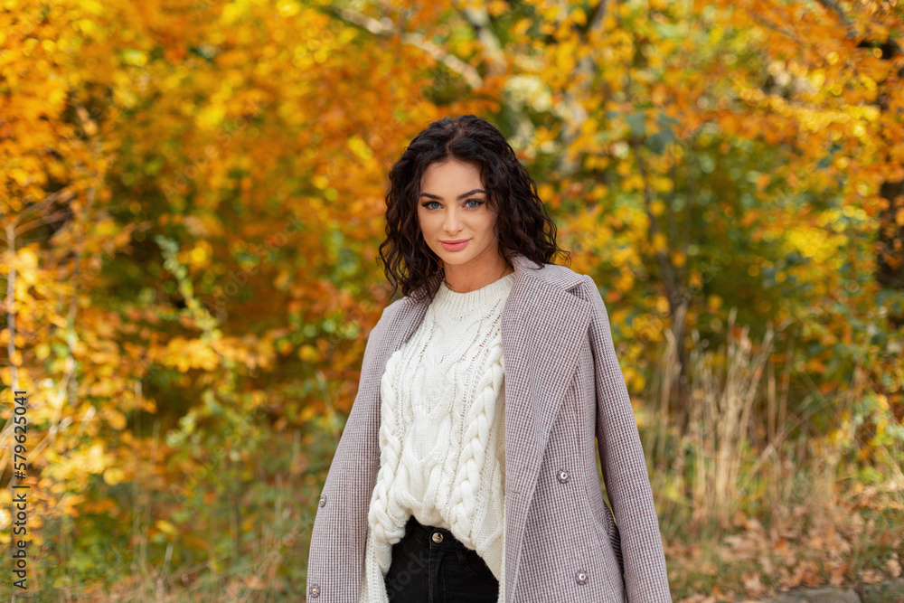 Fashionable beautiful curly girl in a fashion knitted white sweater and coat walks in a golden autumn park against the background of trees Fashionable beautiful curly girl in a fashion knitted white sweater and coat walks in a golden autumn park against the background of trees