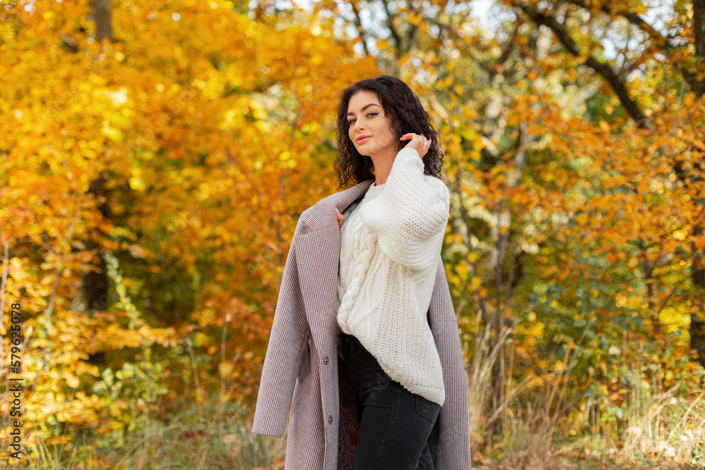 Happy beautiful curly girl model in fashionable autumn clothes with a vintage white sweater and stylish coat walks in a golden autumn park Happy beautiful curly girl model in fashionable autumn clothes with a vintage white sweater and stylish coat walks in a golden autumn park