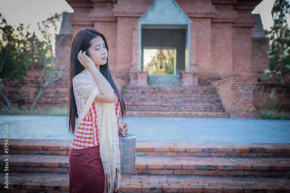 Beautiful thai woman wearing traditional dress standing while holding a tiffin in front of an old temple.