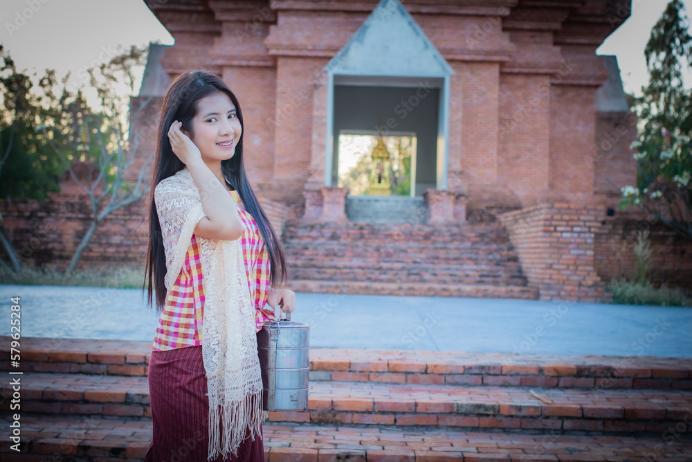Beautiful thai woman wearing traditional dress is standing in front of an old temple holding a tiffin with a smile on her face in the evening.