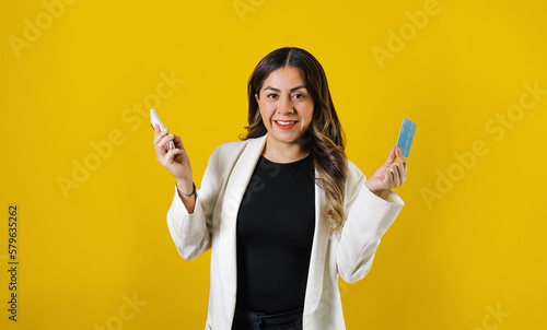 young hispanic business woman using phone and credit card for shopping posing isolated over yellow background in Mexico Latin America
