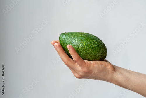 Fresh green ripe avocado in hand of female on light grey background. Seasonal organic raw food for healthy diet, vitamins, vegan, good mood. Vegetable for prepare cooking guacamole on home kitchen