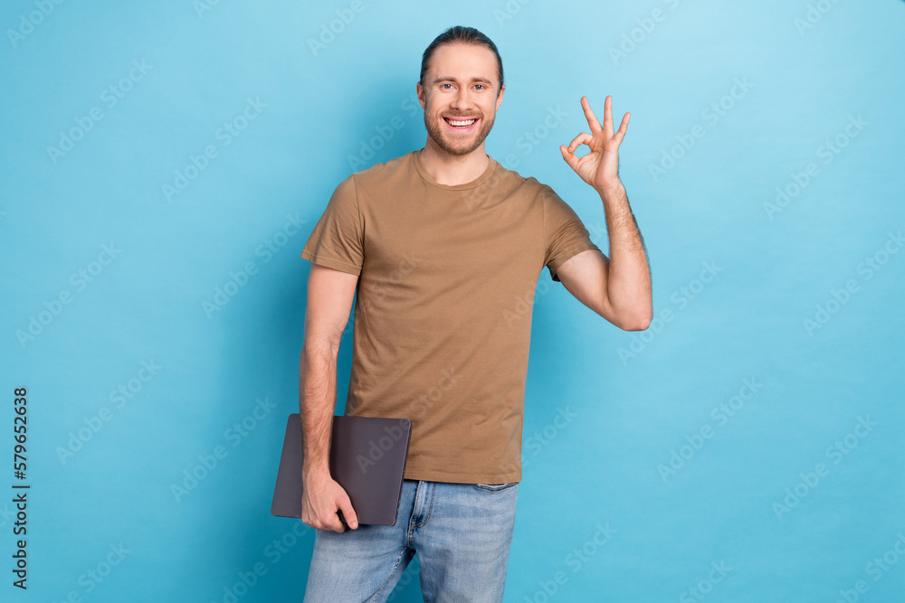 Photo of young guy wear brown t-shirt brunet hair show okey sign hold netbook eshopping advert hp electronics isolated on blue color background Photo of young guy wear brown t-shirt brunet hair show okey sign hold netbook eshopping advert hp electronics isolated on blue color background