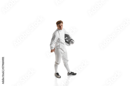 Young man, male fencer in fencing costume mask standing with sword over white studio background. Sport, motivation, professional skills