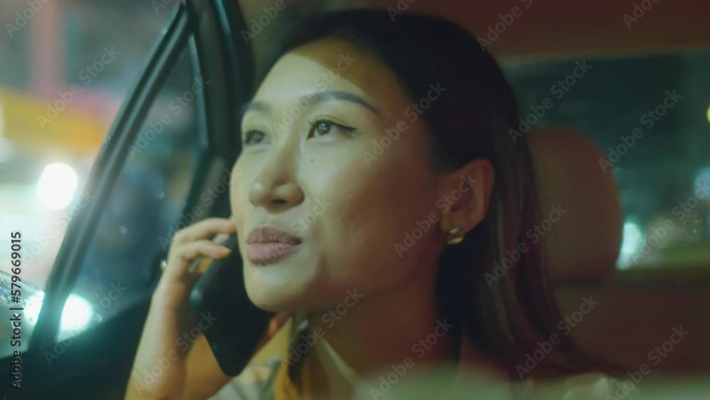 Close up shot of young Asian woman looking at night city through through opened window and speaking on mobile phone during ride Close up shot of young Asian woman looking at night city through through opened window and speaking on mobile phone during ride