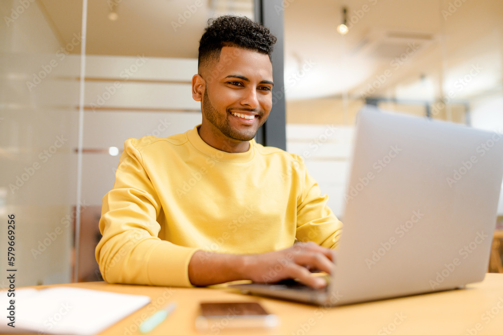 Successful freelancer. Happy millennial Indian man sitting at desk with laptop, smiling and typing, enjoying remote work, male student studying on the distance Successful freelancer. Happy millennial Indian man sitting at desk with laptop, smiling and typing, enjoying remote work, male student studying on the distance