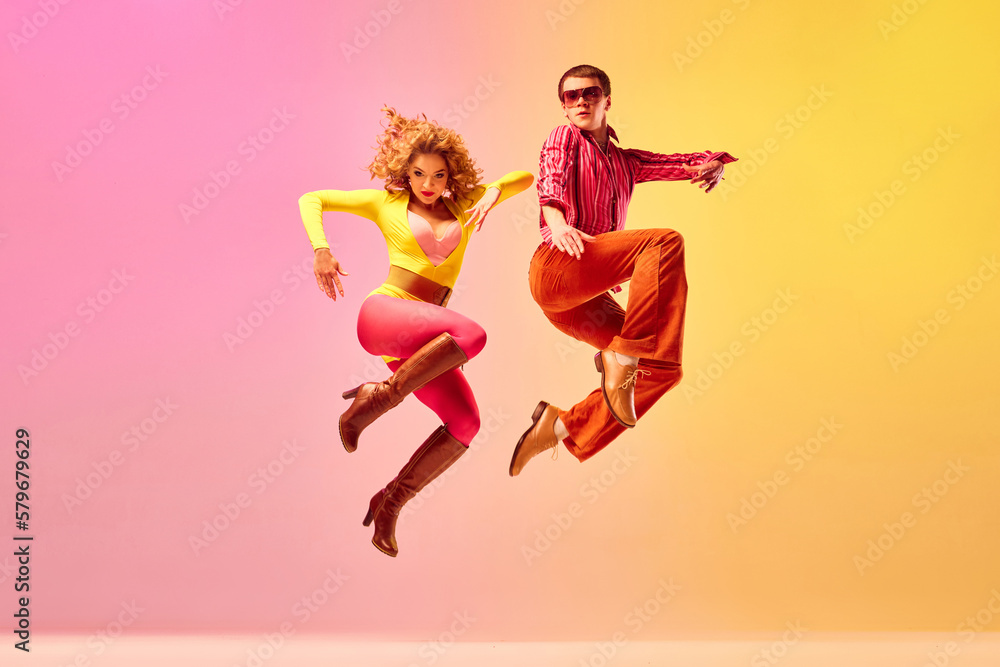 Stylish expressive excited couple of professional dancers in retro style clothes dancing disco dance over pink-yellow background. Concept of 70s, 80s fashion style, music and emotions