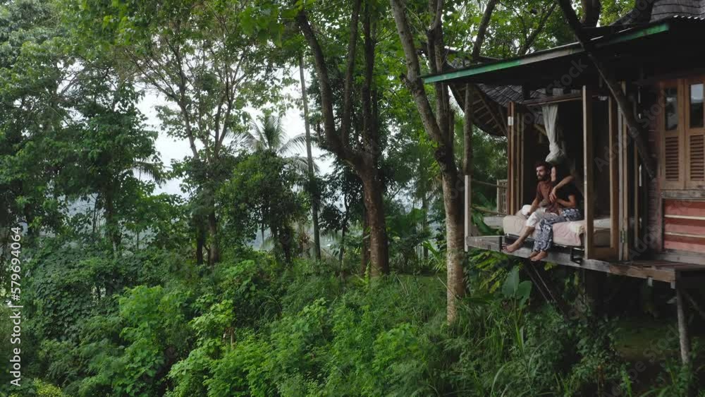 Romantic couple sit by treehouse cabin in forest in Indonesia, aerial pull-out Romantic couple sit by treehouse cabin in forest in Indonesia, aerial pull-out