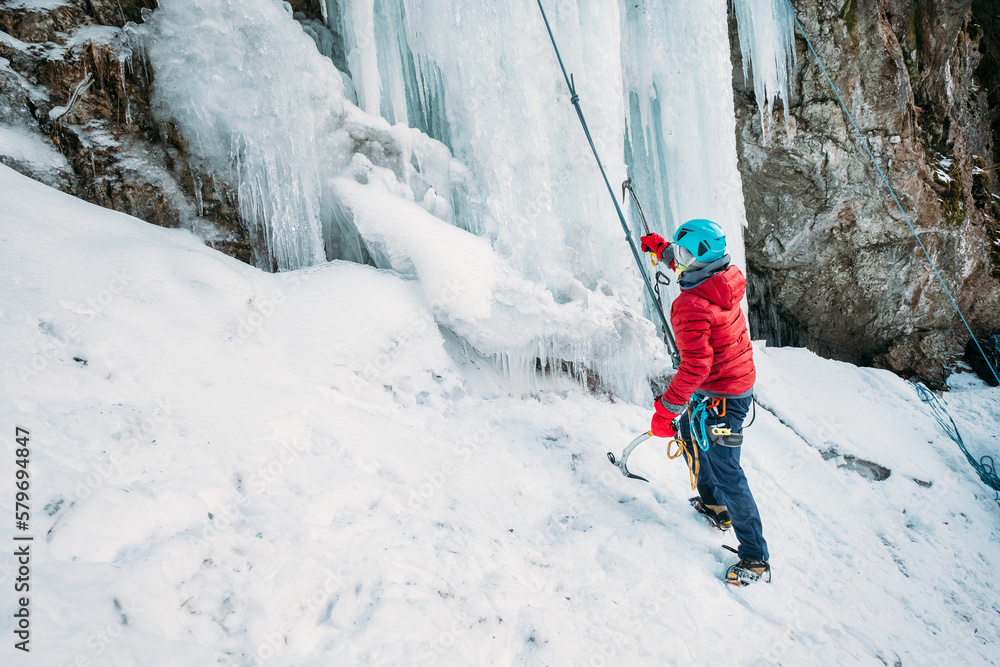Ice climber dressed warm winter climbing clothes, safety harness and helmet under the frozen waterfall. Active people and sporty activities concept image.