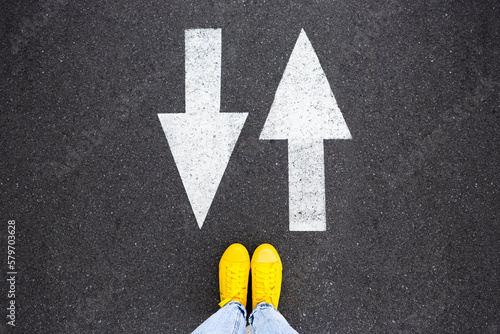 Yellow sneakers on the asphalt road with drawn arrows pointing to two directions. Making decisions and making choices