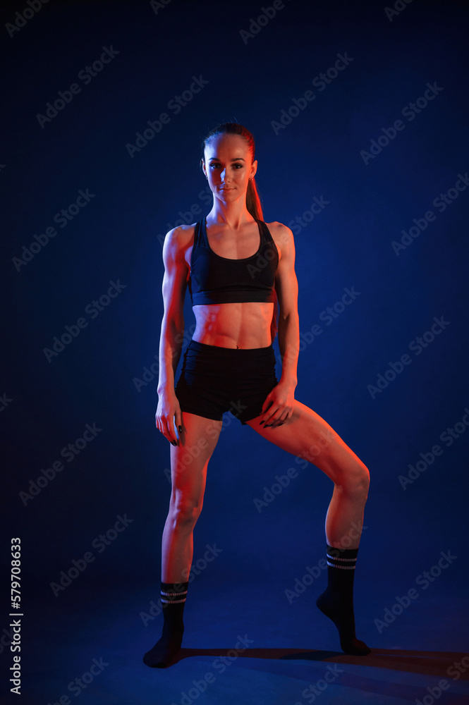 Front view, standing. Beautiful muscular woman is indoors in the studio with neon lighting