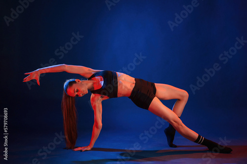 Exercise by standing on a leg and hand. Beautiful muscular woman is indoors in the studio with neon lighting