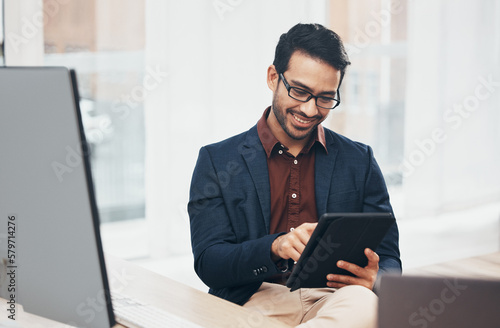 Office, happy Indian man at desk with tablet and computer, reading good news email or successful sales report online. Business, smile and communication with internet, social media or research on web.