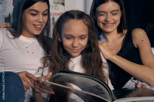 Staged photo. Lesbian couple and their kids are having a good time at home. Cozy atmosphere. The girl is looking at a picture book with her parents. One of the parents is hugging her gently.