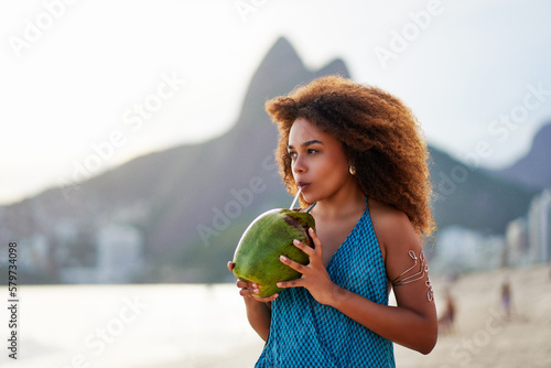 brazilian black young woman drinking coconut water on the beach at sunset in Ipanema, Brazil	