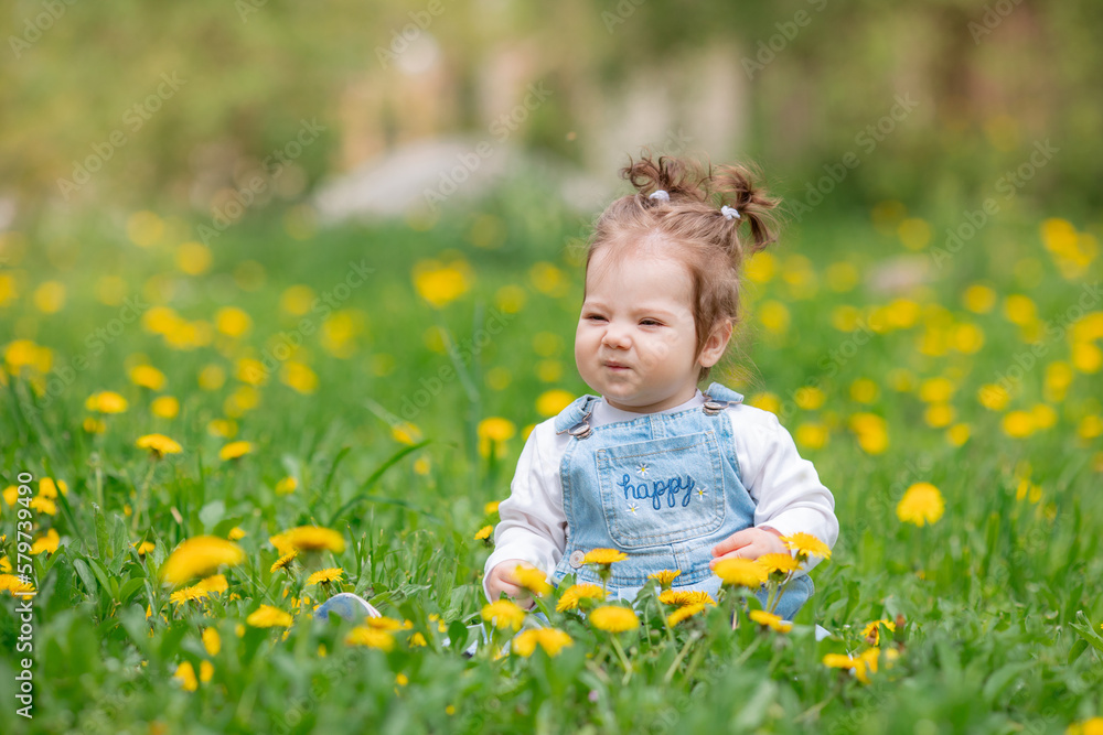 Happy little baby girl sitting on a green meadow with yellow flowers dandelions on the nature in the park Happy little baby girl sitting on a green meadow with yellow flowers dandelions on the nature in the park