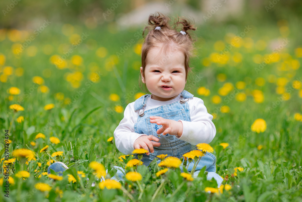 Happy little baby girl sitting on a green meadow with yellow flowers dandelions on the nature in the park Happy little baby girl sitting on a green meadow with yellow flowers dandelions on the nature in the park