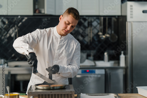 A chef in black gloves flips delicious burger patties on a hot pan with a spatula professional kitchen