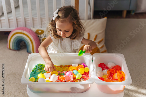 A little girl playing with colored rice and Easter eggs in sensory bin. Easter sensory bin with bright rice and eggs, bunny, carrot. Sensory play and holidays activity for kids. Happy Easter concept. photo