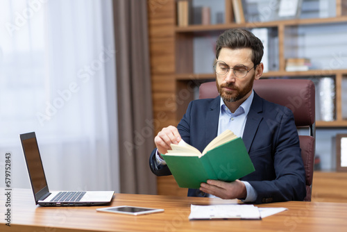 A young man is sitting in the office in a suit at the table, holding and concentrating on reading a green book. Resting, studying, break.
