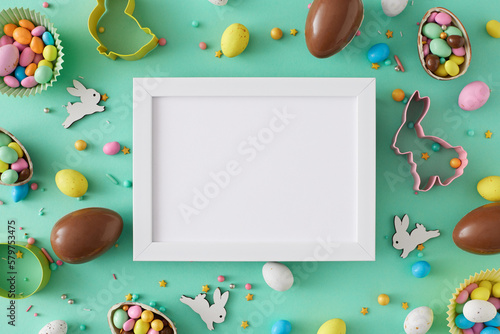 Easter sweets concept. Top view photo of white photo frame chocolate eggs dragees baking molds cute rabbits and sprinkles on teal background with blank space