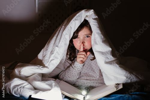 Cute girl reading book under blanket and showing secret gesture photo