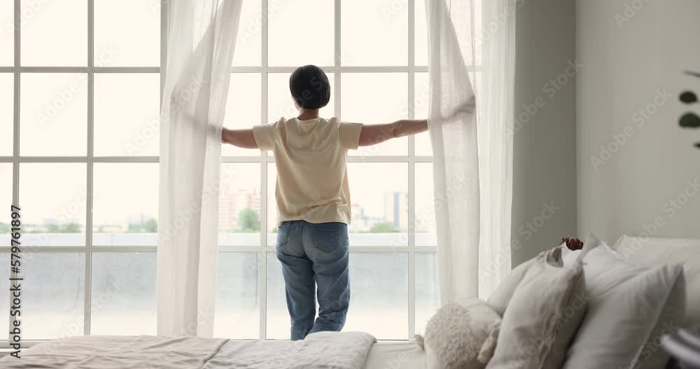 Back view of middle aged homeowner woman parting white transparent curtains in bedroom, opening drapes, looking out of big window at city, enjoying view, natural day light, good morning Back view of middle aged homeowner woman parting white transparent curtains in bedroom, opening drapes, looking out of big window at city, enjoying view, natural day light, good morning