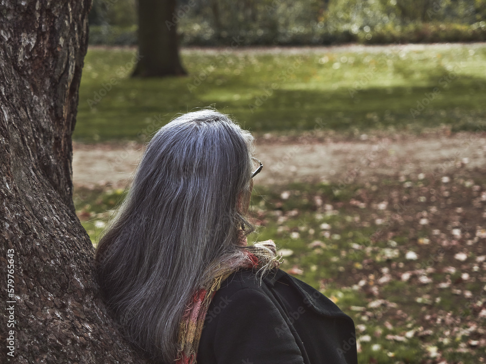 Older woman with gray hair and glasses next to a tree feeling it. Woman leaning on the trunk of a tree in a park with green grass. Girl in prairie and green forest pensive and nostalgic Older woman with gray hair and glasses next to a tree feeling it. Woman leaning on the trunk of a tree in a park with green grass. Girl in prairie and green forest pensive and nostalgic