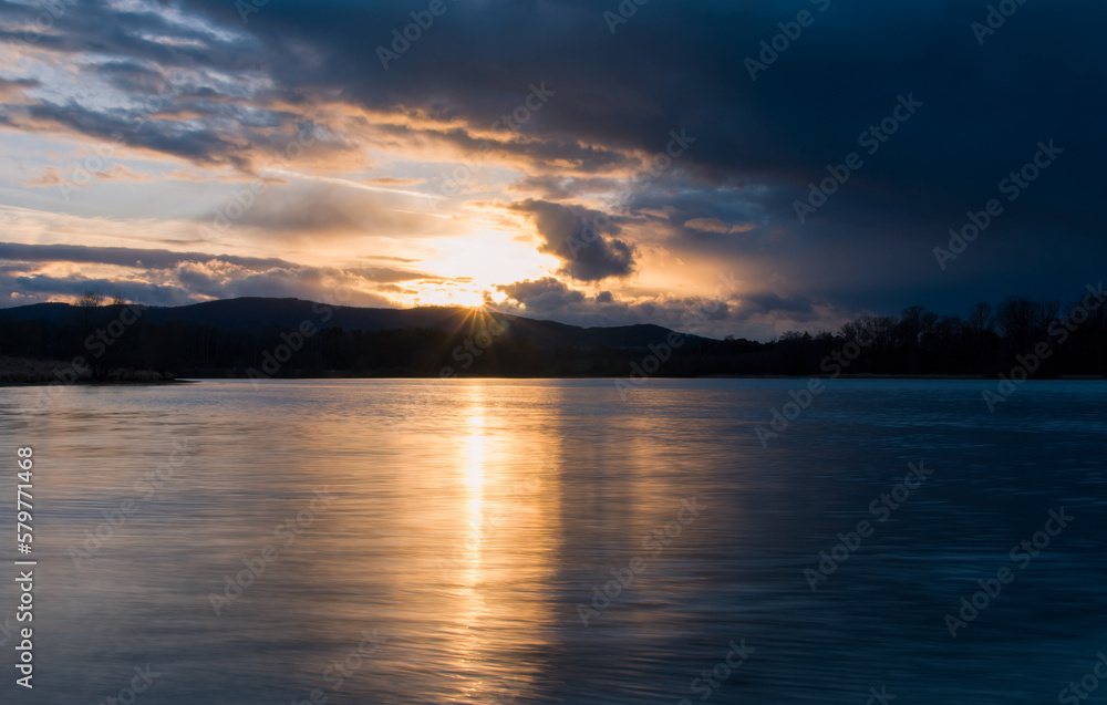 Sunset on pond with distant hill and tree silhouette. Early spring Czech landscape, long exposure