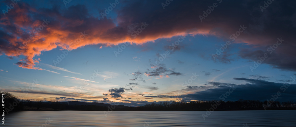 Panoramic sunset sky on pond with distant hill and tree silhouette. Early spring Czech landscape, blue hour, long exposure