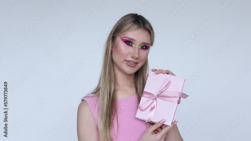 Beautiful young blonde with long hair and neon makeup in pink dress smiles at camera and shows pink gift box with bow. Beautiful young blonde with long hair and neon makeup in pink dress smiles at camera and shows pink gift box with bow.