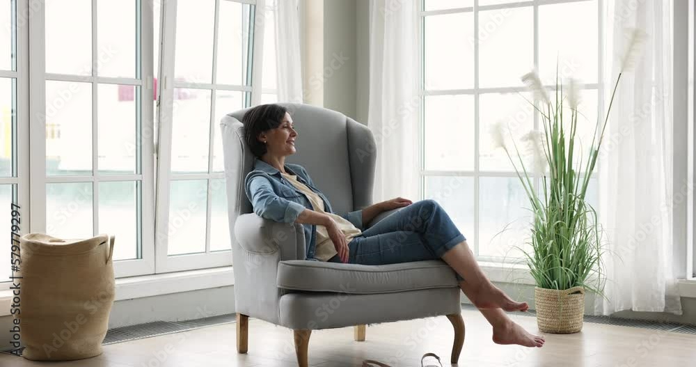 Happy pretty mature lady resting in comfortable home armchair at big windows, listening to relaxing music, enjoying leisure, break, being in cozy warm apartment interior. Side full length shot Happy pretty mature lady resting in comfortable home armchair at big windows, listening to relaxing music, enjoying leisure, break, being in cozy warm apartment interior. Side full length shot