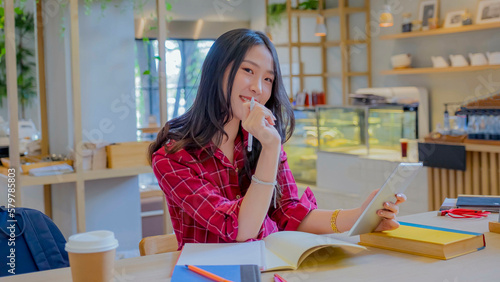 Young smiling asian woman with long brown hair, dressed in a red white striped shirt, is holding a tablet computer and a pen at a study desk. beautiful sitting at cafe she relaxed. out of focus. photo