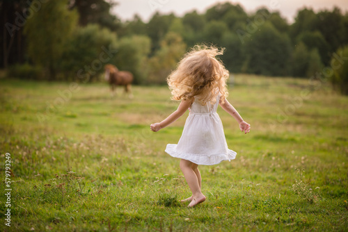A little girl with long hair runs barefoot on the ground. Beautiful child playing at sunset in the forest in summer