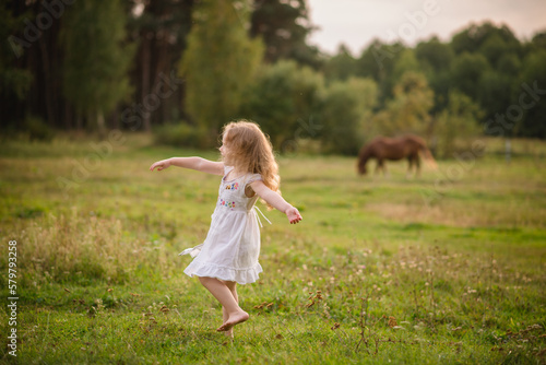 A little girl with long hair runs barefoot on the ground. Beautiful child playing at sunset in the forest in summer