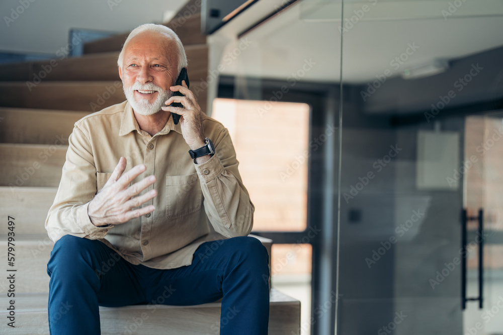 Happy relaxed senior man wearing beige shirt and hand watch talking on mobile phone while sitting on stairs at home during the day. Technology, people, lifestyle and communication concept Happy relaxed senior man wearing beige shirt and hand watch talking on mobile phone while sitting on stairs at home during the day. Technology, people, lifestyle and communication concept