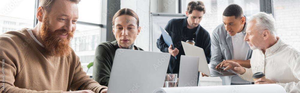 Positive businessman using laptop near tattooed colleague in office, banner. Positive businessman using laptop near tattooed colleague in office, banner.