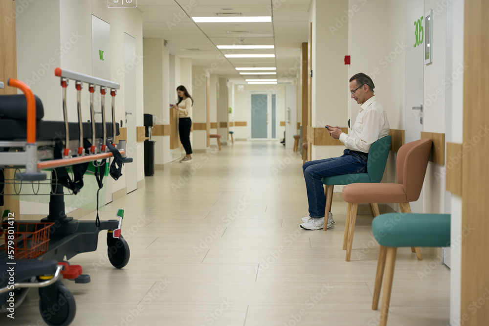 Fototapeta premium Man in glasses sits in a hospital corridor with a phone in his hands