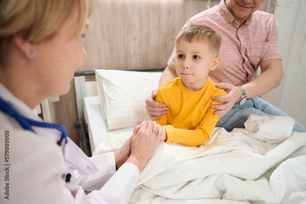 Neat child on the medical bed is looking to the doctor in the ward Neat child on the medical bed is looking to the doctor in the ward