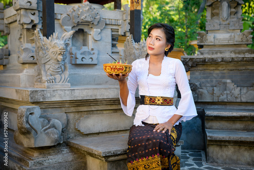 Portrait of beautiful Asian Bali Woman with beauty face and attractive full lips wearing a white shirt and brown sarong holding offerings photo