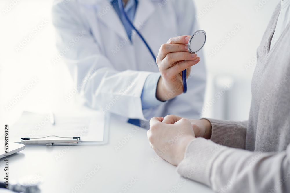 Doctor and patient sitting at the table in clinic. The focus is on female physician's hands examining woman with stethoscope, close up. Medicine concept. Doctor and patient sitting at the table in clinic. The focus is on female physician's hands examining woman with stethoscope, close up. Medicine concept.