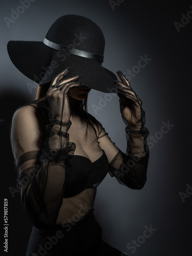 Fototapeta Naklejka Na Ścianę i Meble -  Fashion portrait of a young girl in a black bra and transparent top. Felt black hat. Model posing, faces not visible photo in studio.