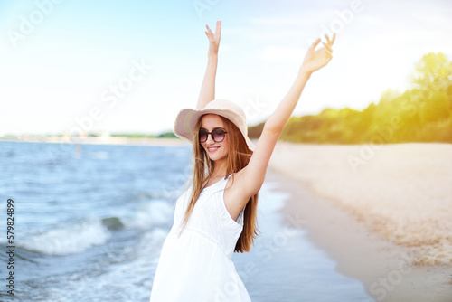 Happy smiling woman in free happiness bliss on ocean beach standing with a hat  sunglasses  and rasing hands. Portrait of a multicultural female model in white summer dress enjoying nature