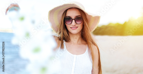 Happy smiling woman in free happiness bliss on ocean beach standing with hat, sunglasses, and white flowers. Portrait of a multicultural female model in white summer dress enjoying nature © rogerphoto